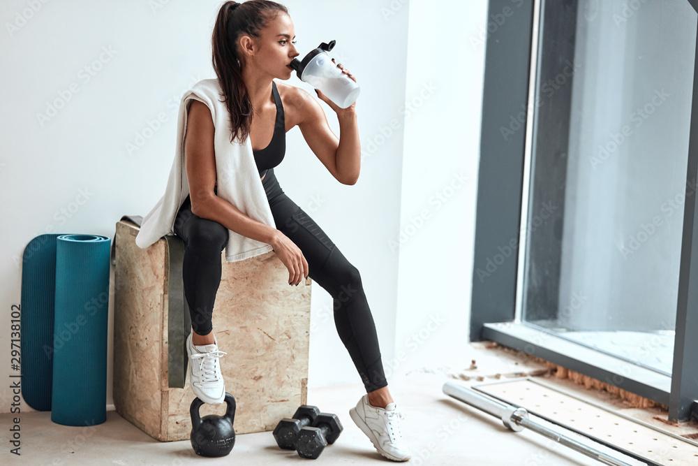 Young woman in leggins with towel on shoulders drinking water after fitness training